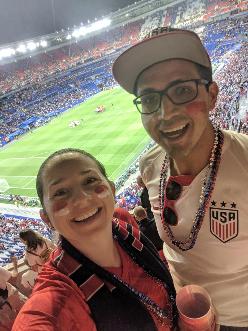 Mark and Lindsay Rojas stand in the stands of a major stadium as a soccer team plays on the field below.