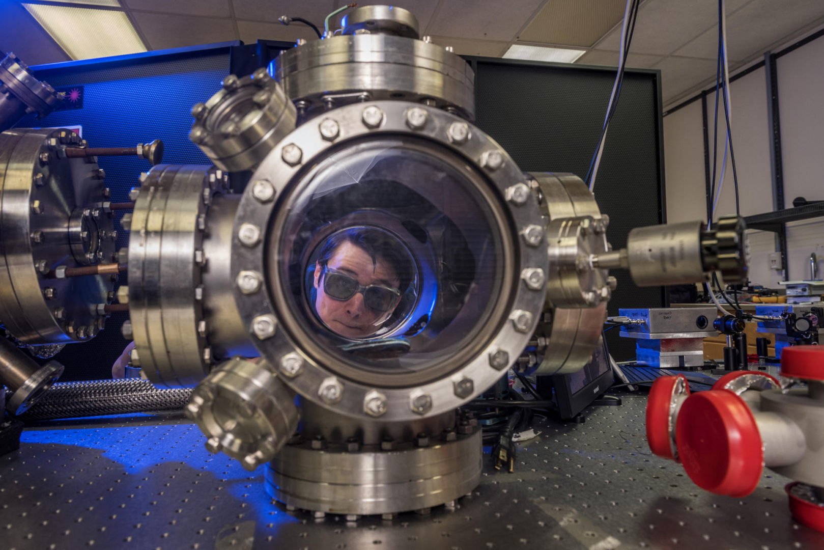 A student wearing protective glasses peers through the clear core of an extensive piece of physics equipment. 