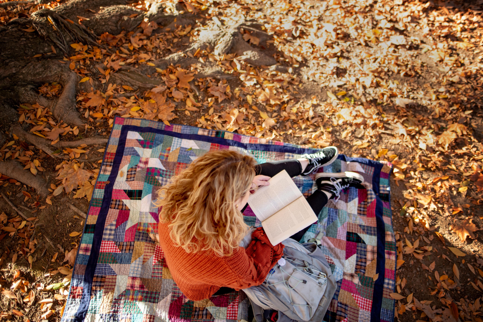 A student sits on a quilted blanket as she reads a book that is opened on her lap.