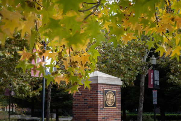 A brick pillar with a University seal stands out amongst trees and leaves changing colors.