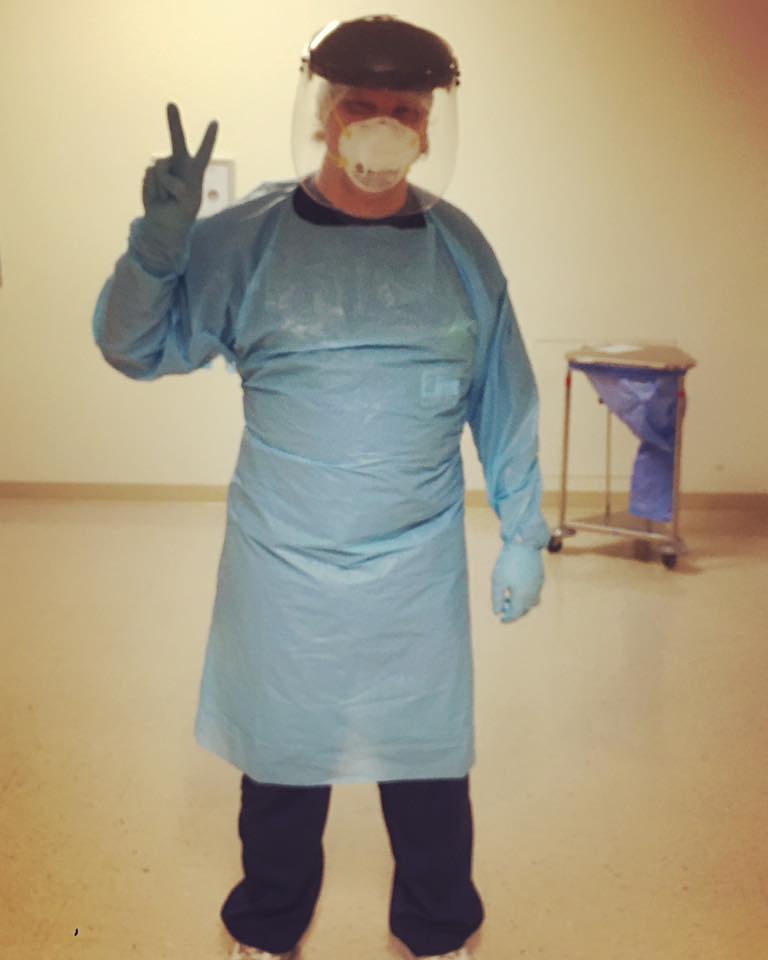 Jared Axen flashes the peace sign with his hand while dressed in scrubs, a protective gown, rubber gloves, a face mask, and a face shield.