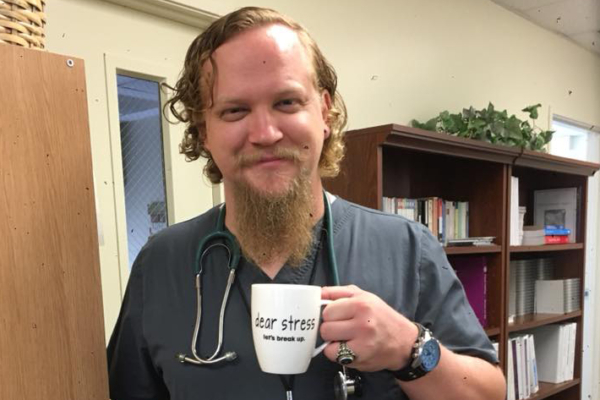 Jared Axen holds a mug while dressed in his scrubs and a stethoscope around his neck.