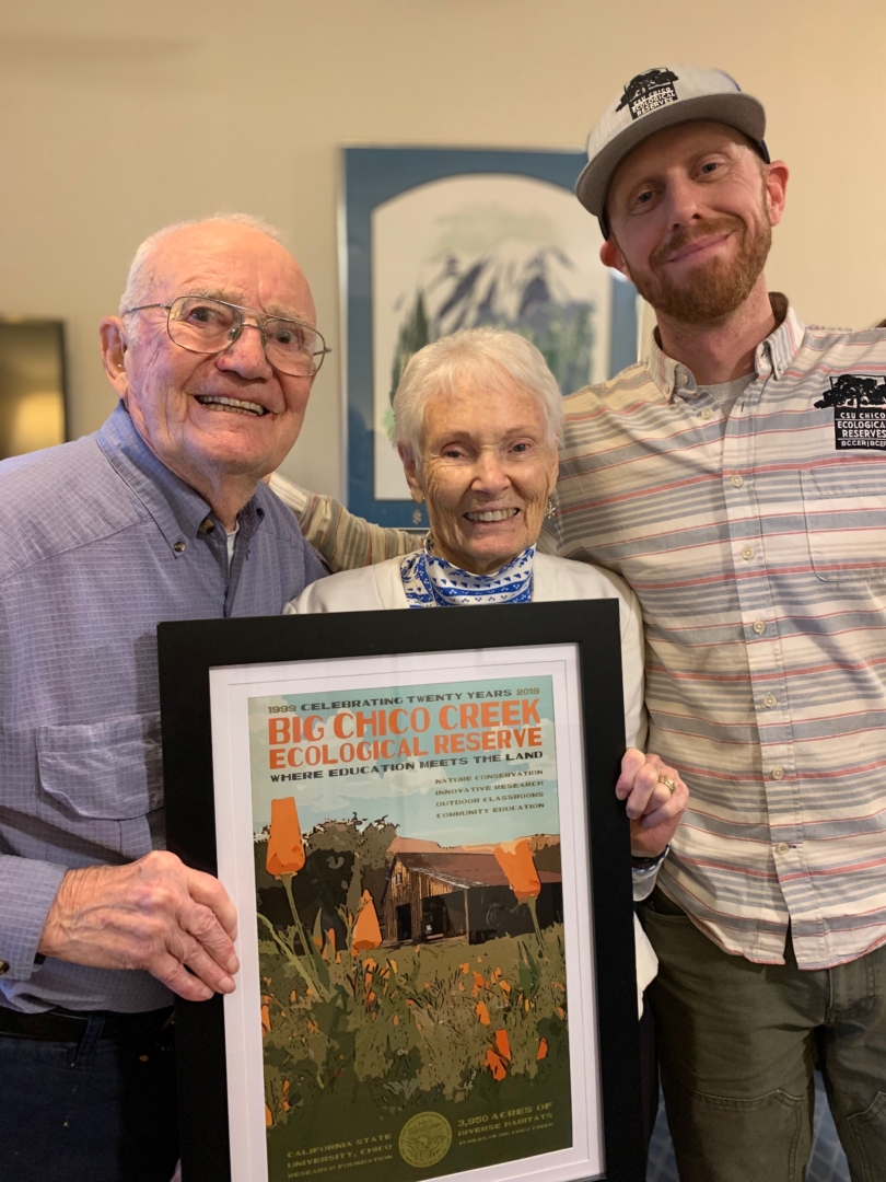 Wes and Phyllis Dempsey pose and smile with Eli Goodsell while displaying a framed poster of the BCCER.