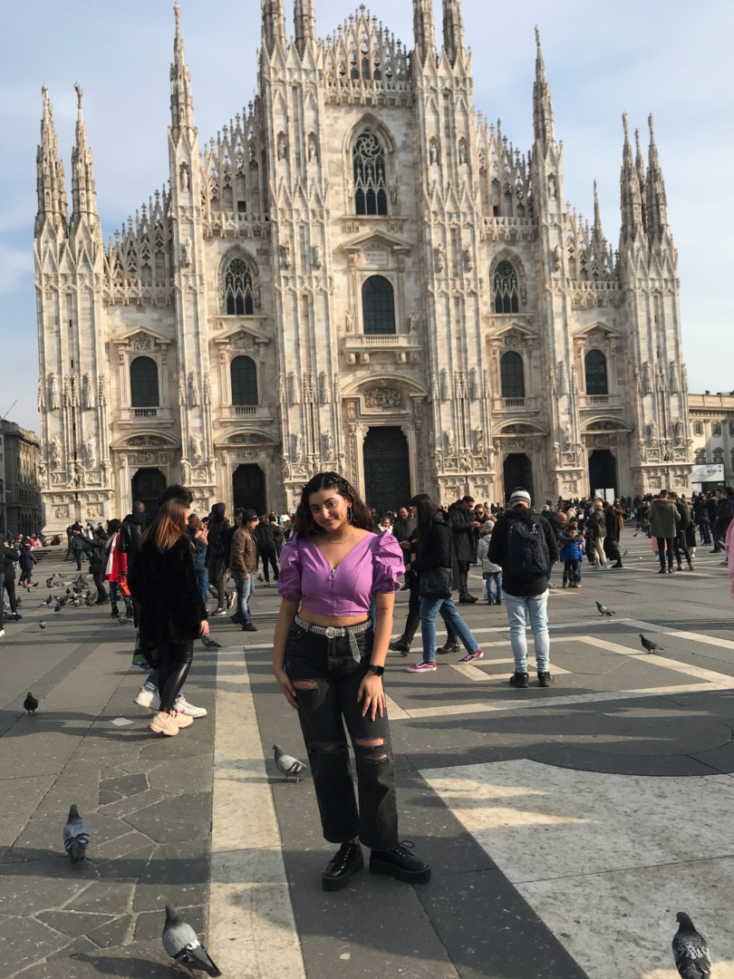 An American college student stands in front of a beautiful building in Italy while tourists mill around in the background.