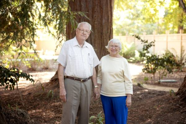 Tom and Melaine Taylor hold hands in front of a redwood tree on campus.