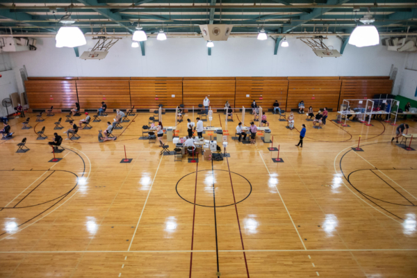 Students and healthcare workers sit in chairs inside a basketball gym.