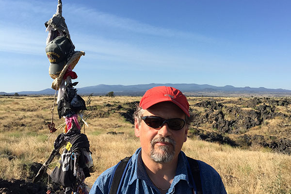Jesse Dizard stands on an open ridge in the foothills in front of a wooden pole with clothes and hats hanging from it.