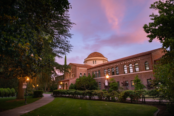 An academic building sits in the foreground as clouds paint the sky during a sunset in the background