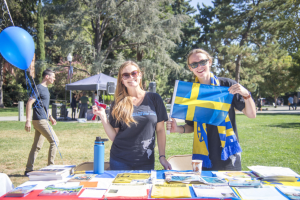 Two students, one holding a sticker and the other holding a flag from Sweden, stand behind a table with papers and pamphlets.