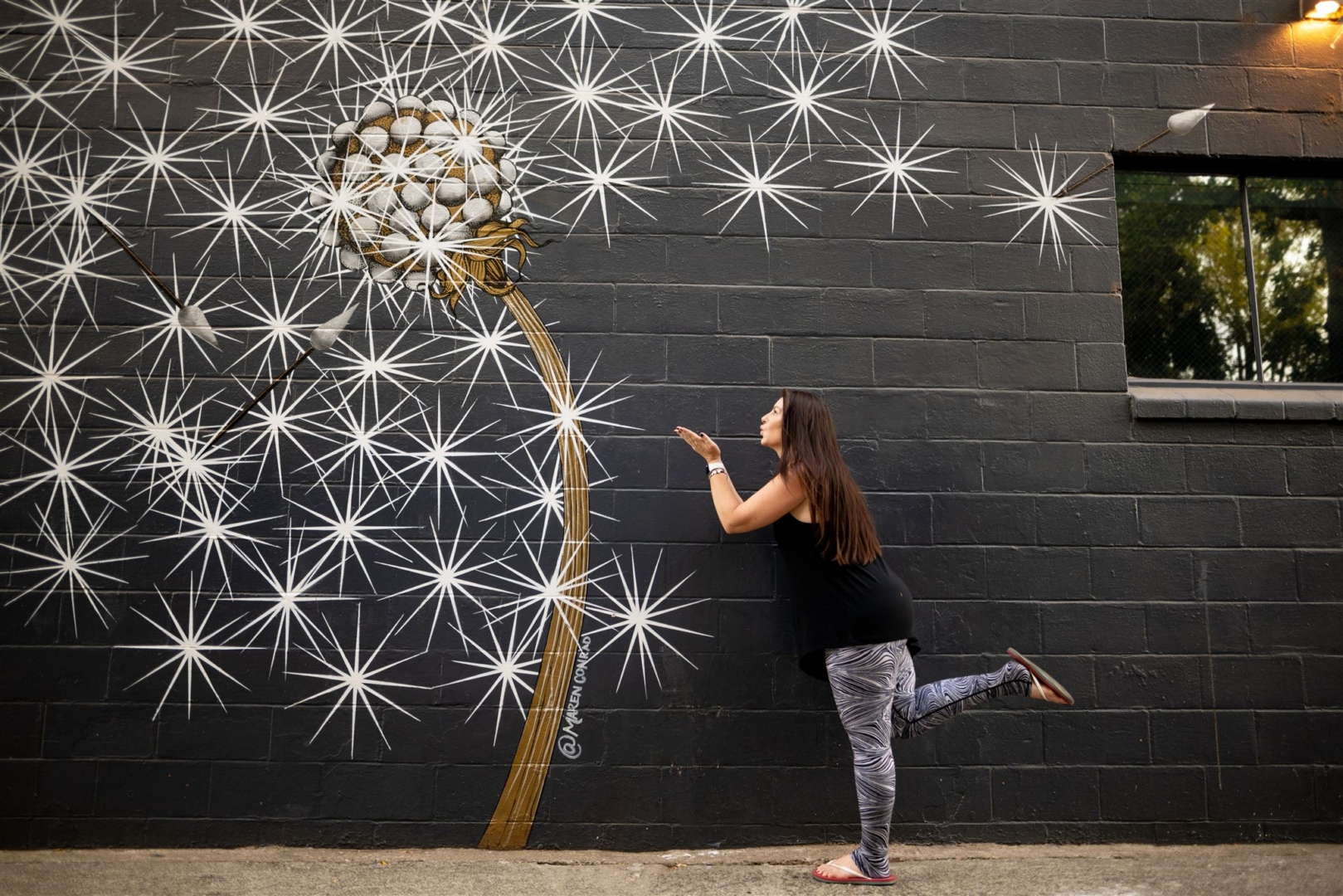 Maren Conrad interacts with her painted mural of daffodils by blowing in the direction of it so it seems she's blowing the daffodil.