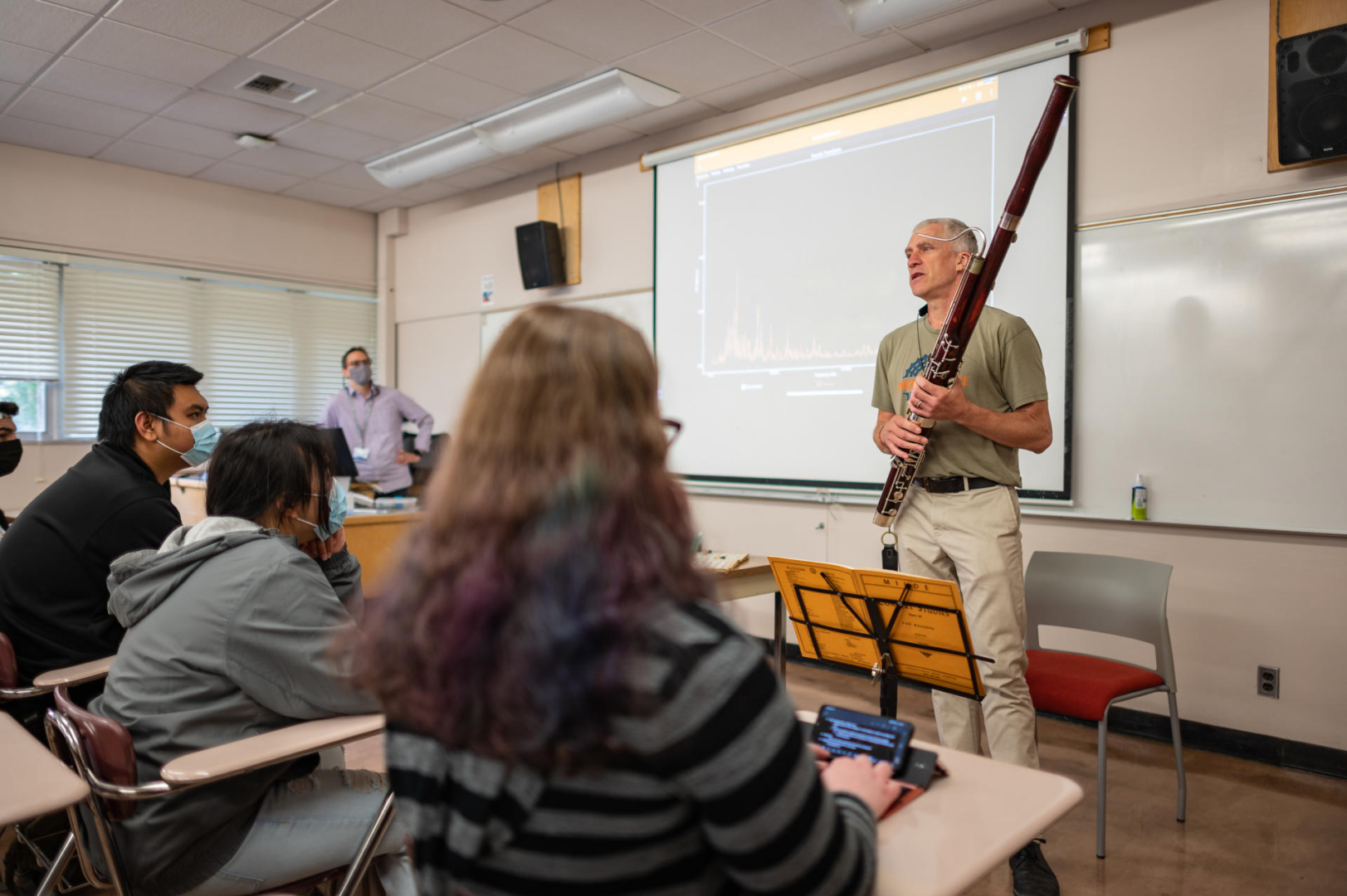 A man holds a bassoon in front of a classroom of college students.