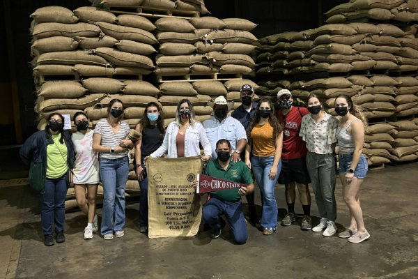 College students and faculty stand in a coffee processing plant, with dozens of bags of coffee beans stacked up in the background.
