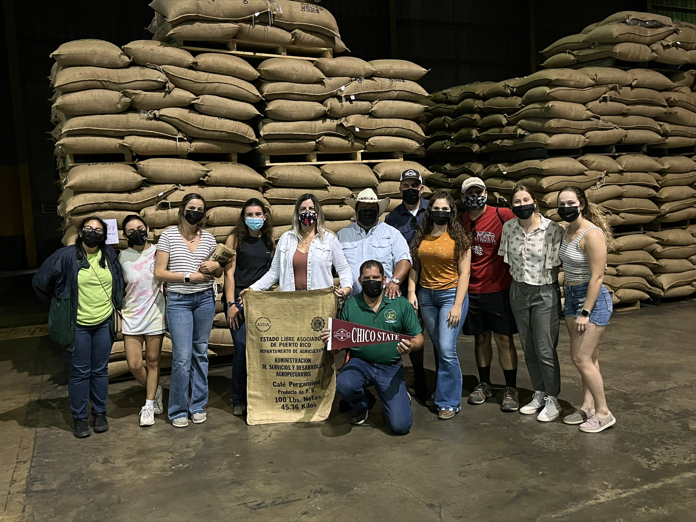 College students and faculty stand in a coffee processing plant, with dozens of bags of coffee beans stacked up in the background.