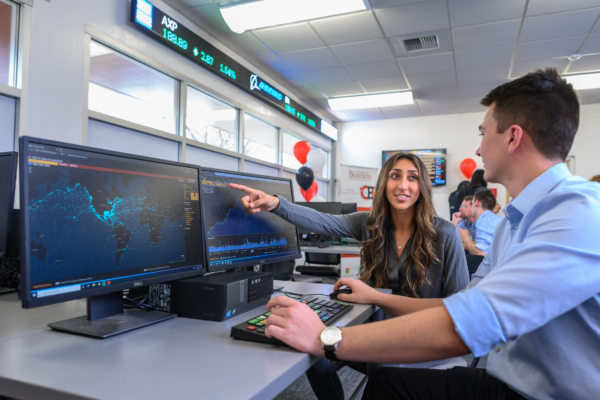 Business students work at a computer with business tickers displayed above them.