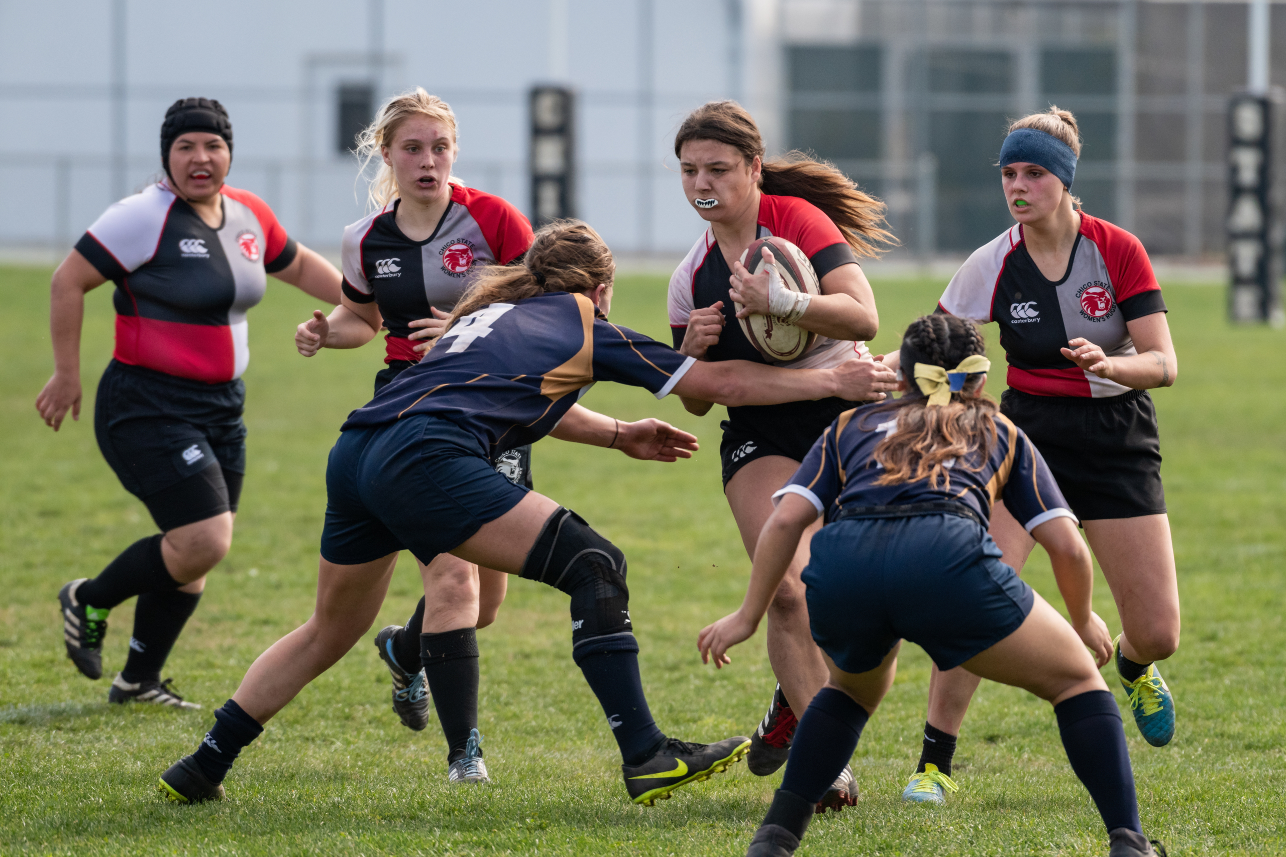 A Chico State Rugby player carries the ball while opposing players attempt to tackle her.