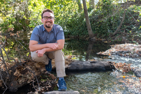 Cole Schmitt, a white cisgender male student, is perched on a log in the creek that runs through Chico State campus.