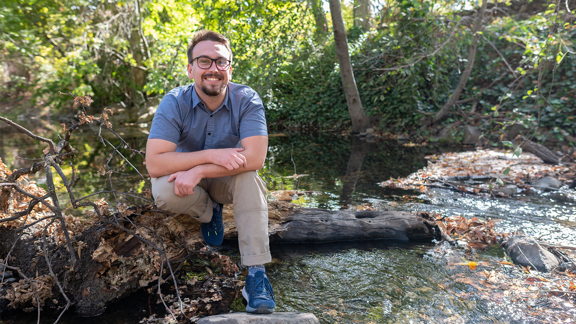 Cole Schmitt, a white cisgender male student, is perched on a log in the creek that runs through Chico State campus.
