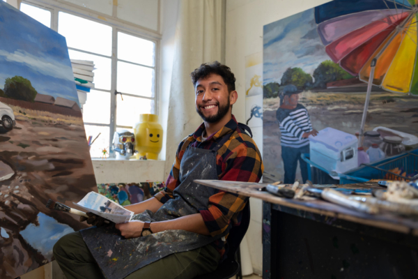 Joel Solis sits and smiles for the camera while holding a photograph he took. His painting of a Latina street vendor is seen in the background.