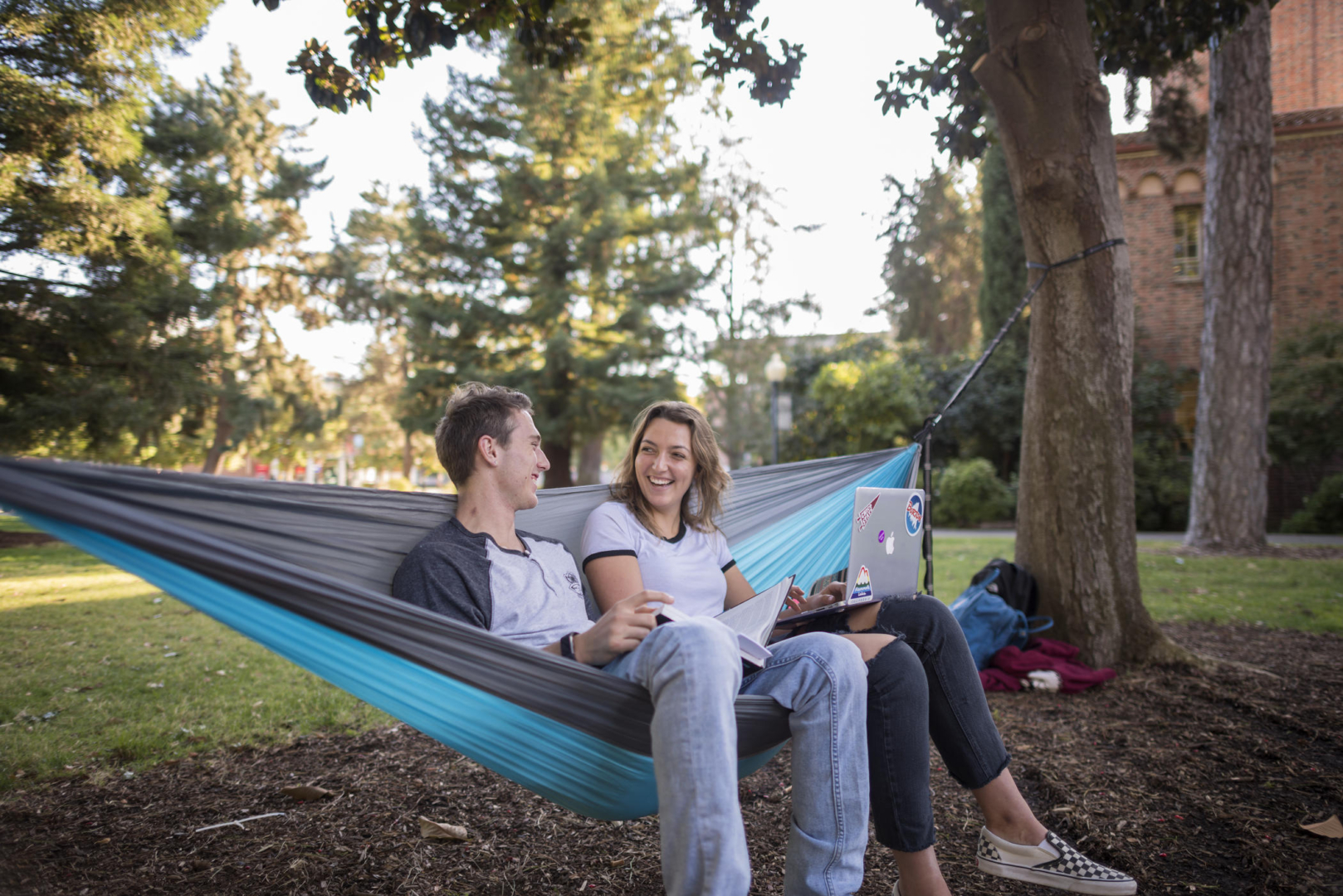 Two students sit on a hammock while studying. One student has a laptop on her lap while the other student holds a textbook.