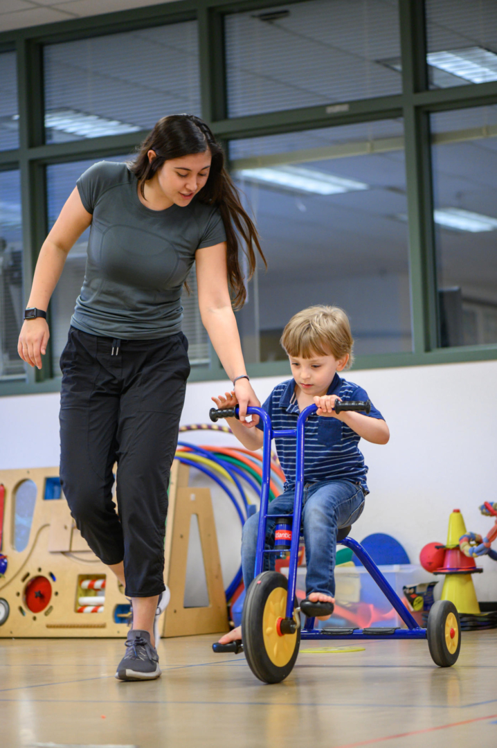 Student walks next to 5-year-old boy riding a tricycle during a session at Chico State's Autism Clinic. 