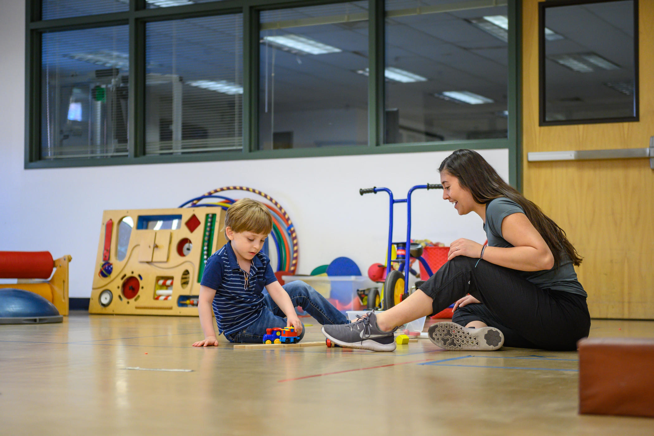 Student sits on the floor with 5-year-old boy as they play with trains during a session in the Autism Clinic in March 2020. Other toys and play equipment, like hula hoops, can be seen in the background.