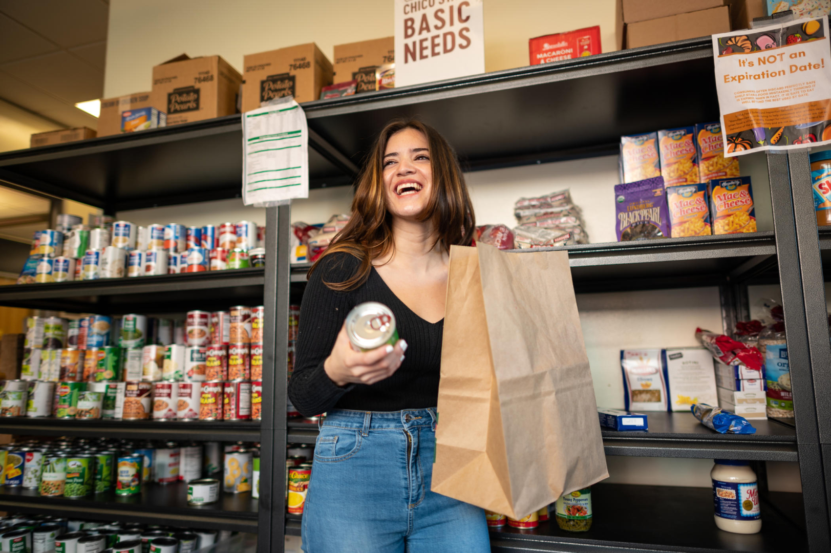 Student holds a paper bag on the left and a can on the right as she stands in front of shelves with non-perishable foods inside the Wildcat Food Pantry.