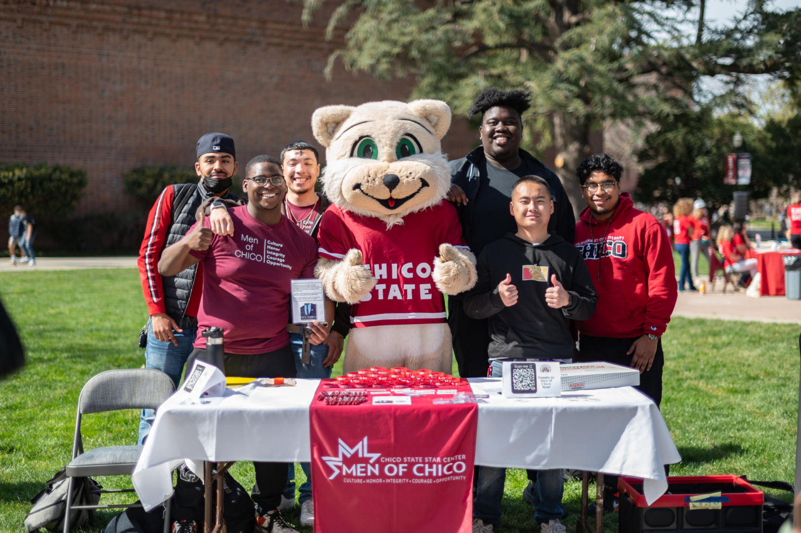 A group of students stands behind a table with a banner that reads "Men of Chico" while they pose for a photo with the Chico State mascot Willie the Wildcat.