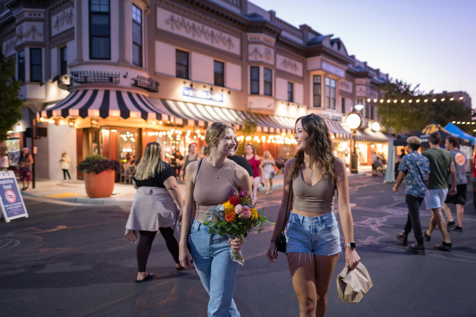 Two students walking in the street during an evening farmers market. One students hold a bouquet of colorful flowers in one hand and the other student holds a paper bag.