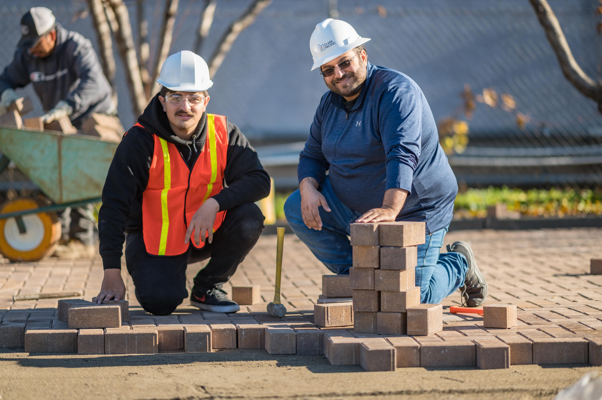 A student (left), wearing an orange safety vest, kneels alongside Professor Mohammed Albahttiti (right) while the two work on installing pavers at the site of a community service project at the Chico Islamic Center.