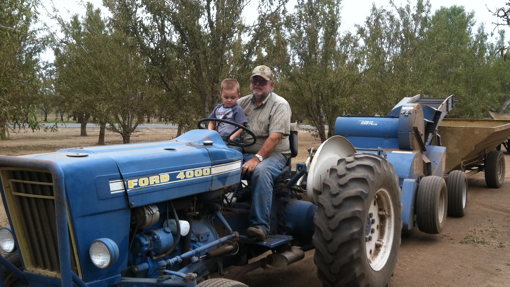 Jerry Miguel and his grandson on a blue tractor in an orchard.