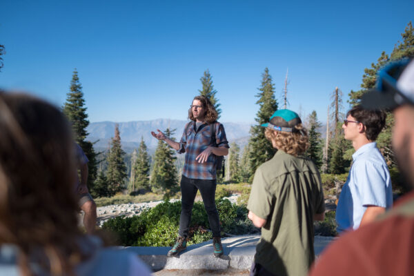 Professor Jesse Engebretson is standing on a rock overlooking a valley of trees. He is addressing a group of students during a field trip.