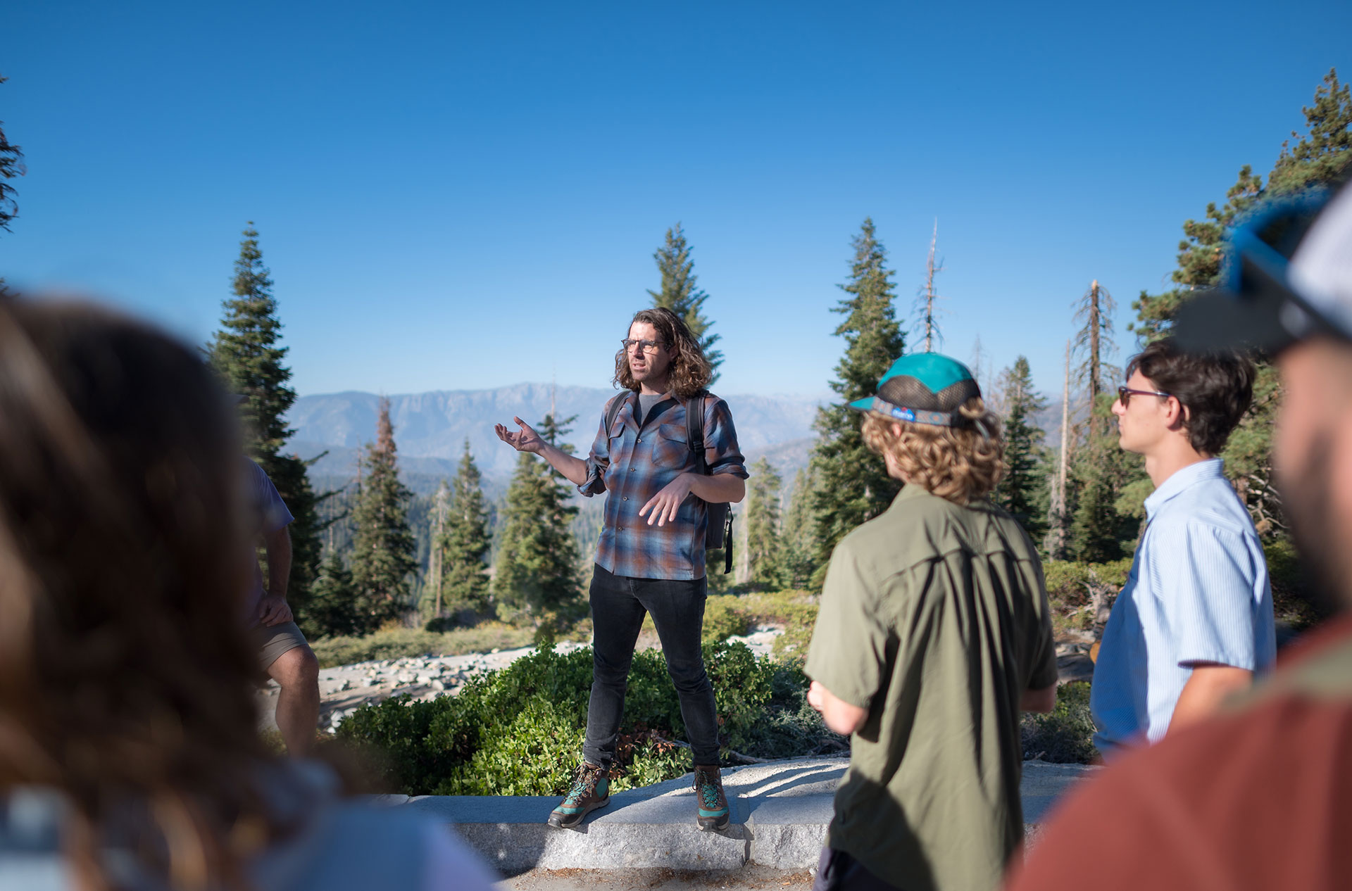 Professor Jesse Engebretson is standing on a rock overlooking a valley of trees. He is addressing a group of students during a field trip.