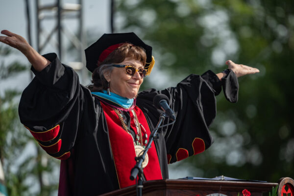 Standing in full regalia on stage, President Hutchinson extends her arms out toward the audience at a Commencement ceremony.