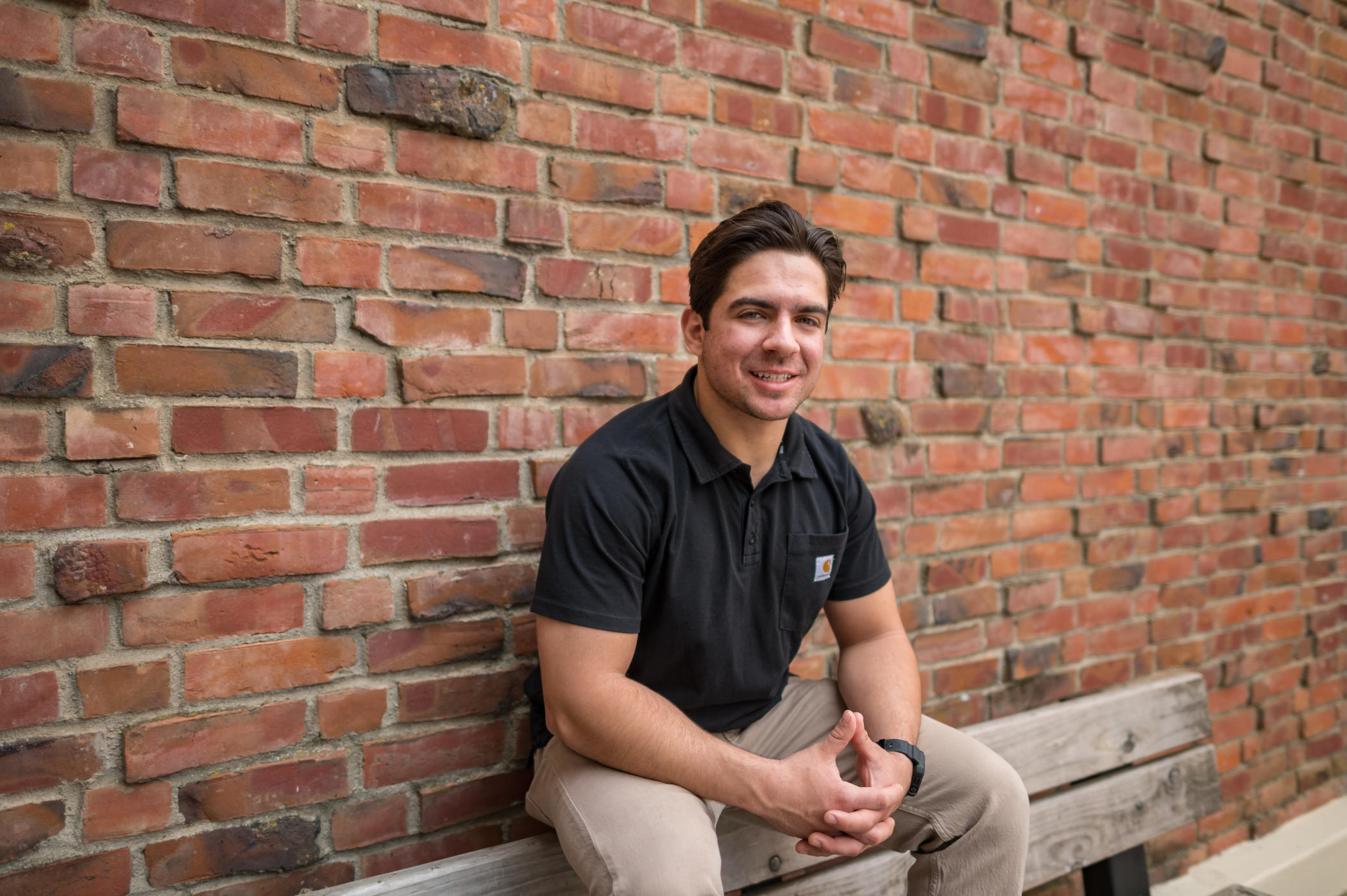 Mateus Avalos, wearing a black polo shirt and khaki pants, sits on a bench in front of a brick building for a posed photo.