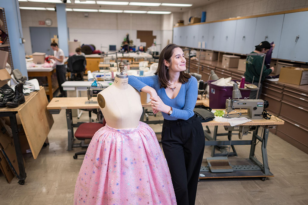 Gaby Saxon poses next to the pink, intricately beaded skirt she's been working on in the costume shop to be worn by the actor playing Glinda "Good Witch of the North" for an upcoming performance of Wizard of Oz.