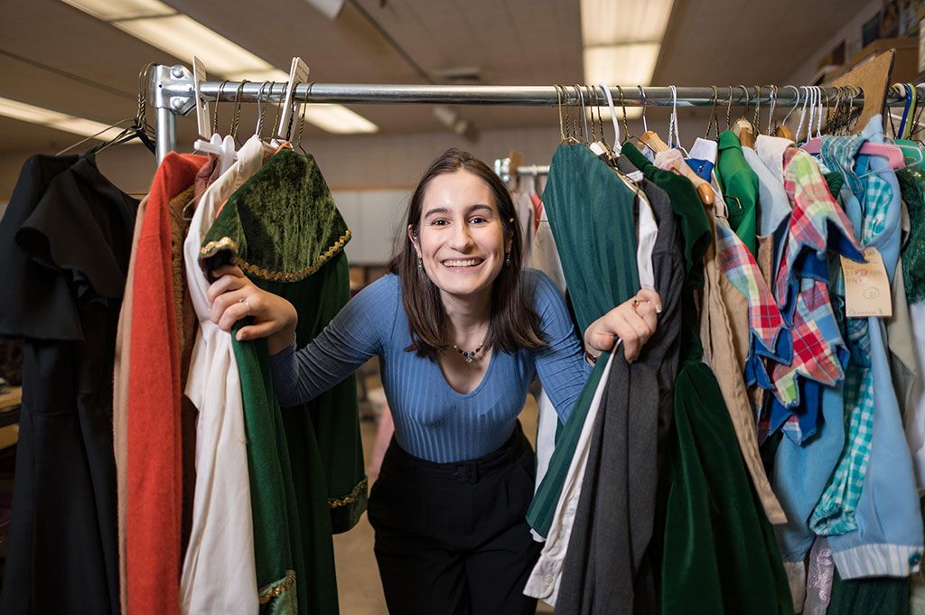 Gaby Saxon poses in the costume shop, appearing in the middle of a rack of hanging costumes.