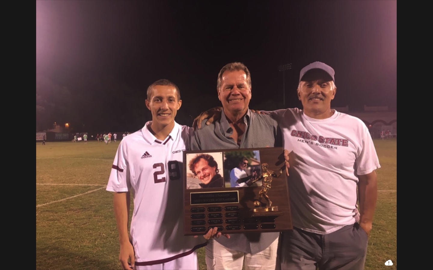 Three men pose for a photo on a soccer field, while the man in the middle holds a memorial plaque in his hands.