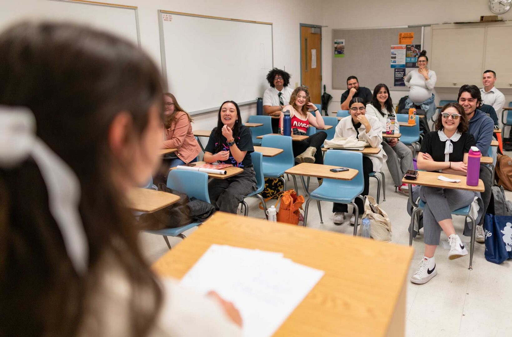 Students in a college class sit at individual desks listening to and smiling at another student.