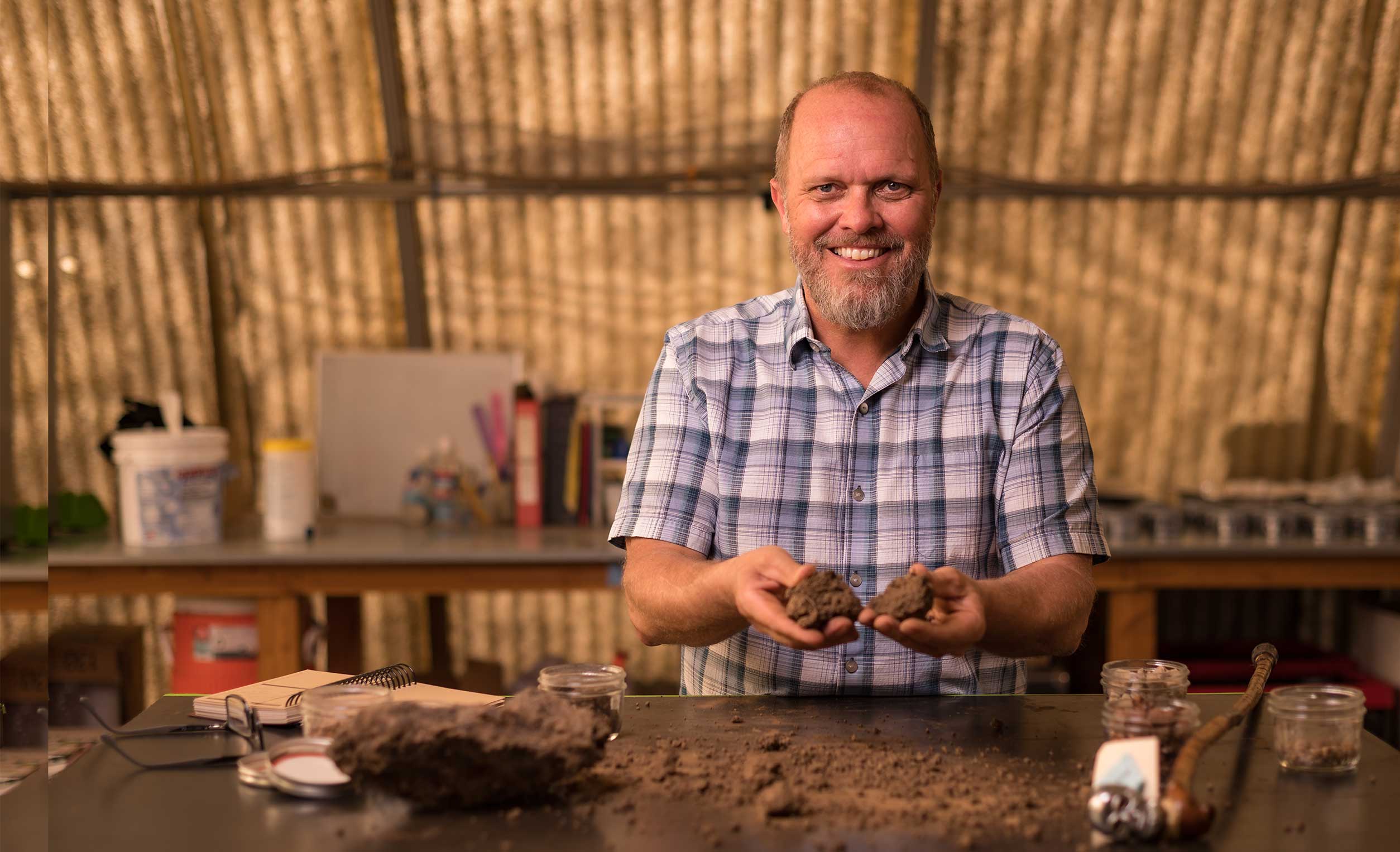 Professor Garrett Liles holds samples of soil in each hand. He is standing at a table inside the soil sampling area of the regenerative agriculture lab.