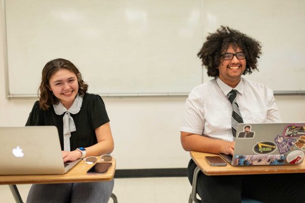 Marin Spalding, left, and Marcus Peoples, on the right, sit side-by-side in a classroom with laptops opened in front of them. Both are dressed in formal clothing, smiling widely and clearly amused.