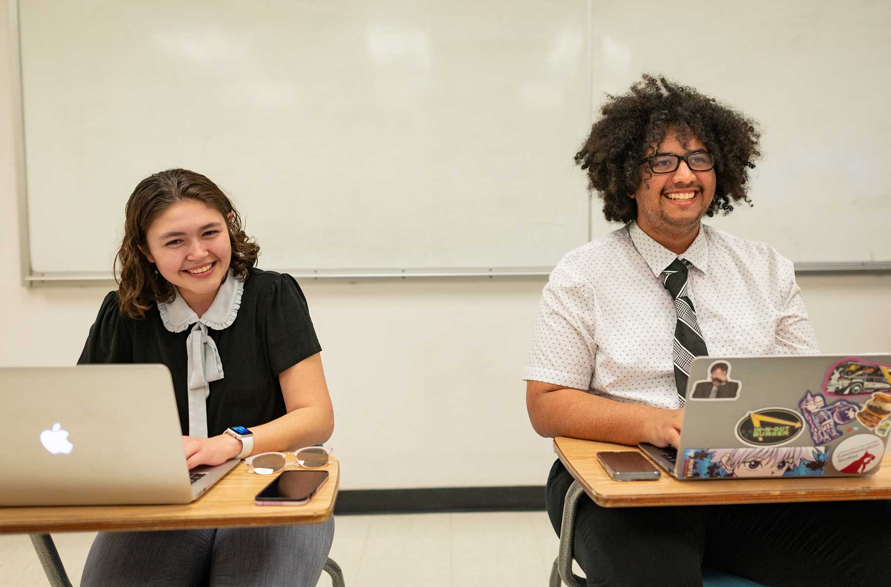 Marin Spalding, left, and Marcus Peoples, on the right, sit side-by-side in a classroom with laptops opened in front of them. Both are dressed in formal clothing, smiling widely and clearly amused.