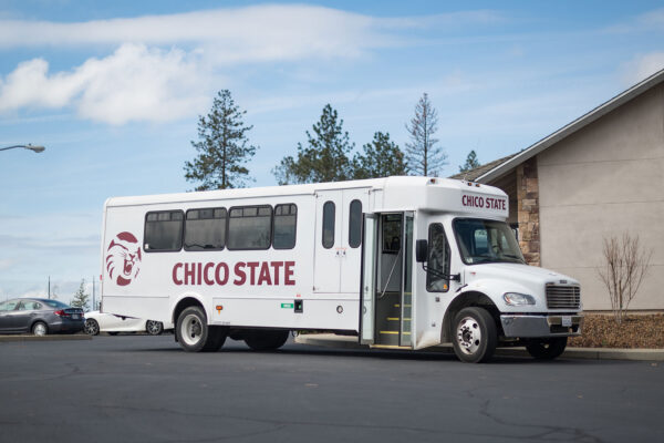 A white bus is parked with its door open; it has Chico State written on the side and the Wildcat logo printed on it