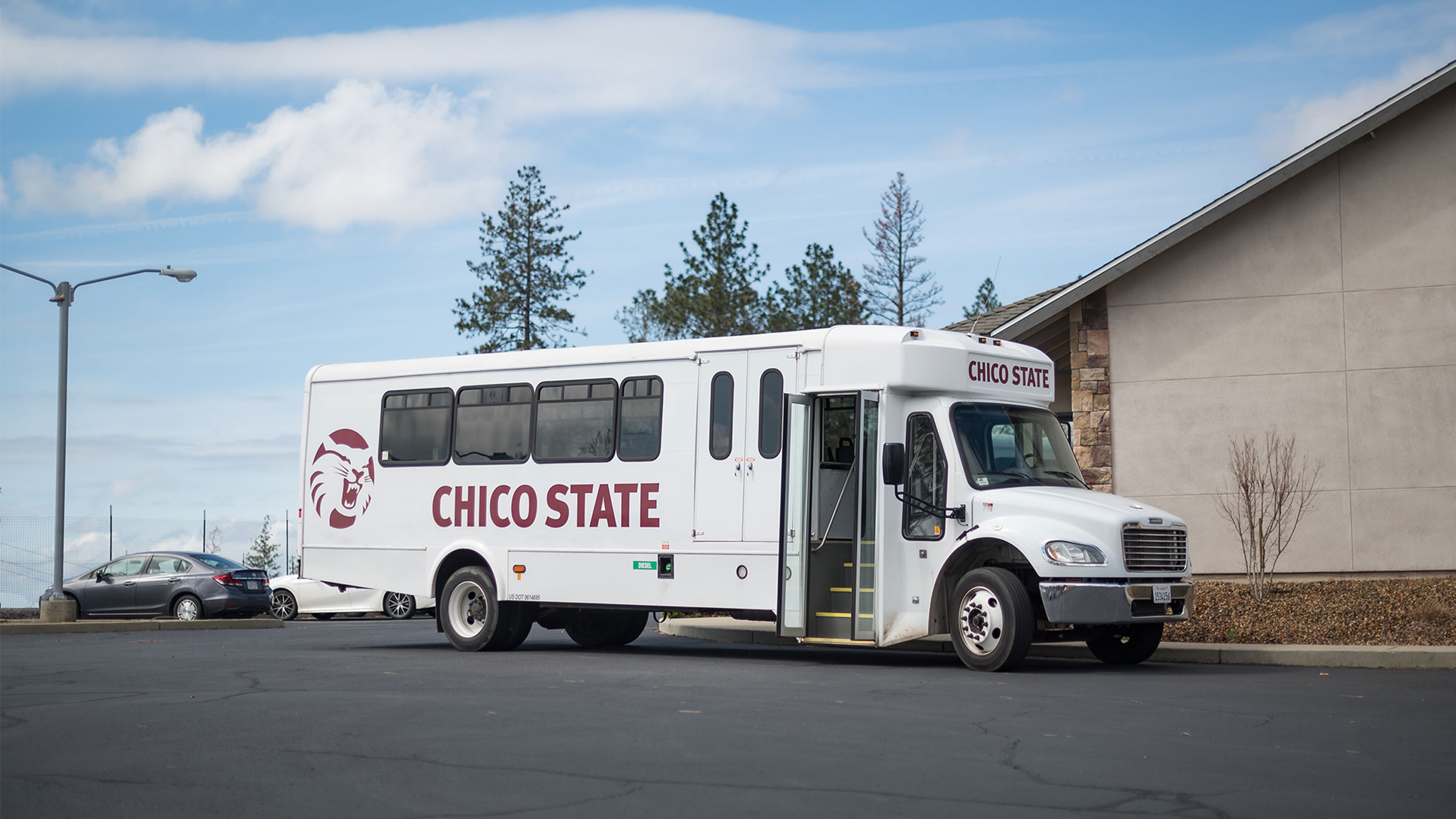 A white bus is parked with its door open; it has Chico State written on the side and the Wildcat logo printed on it