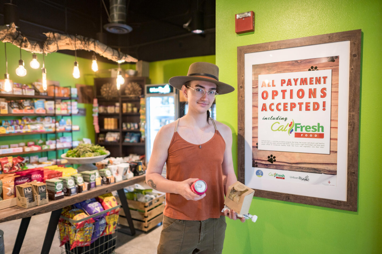 Isa Cooks holds a drink and food items in hands while standing at the entrance of a small market and next to a poster that reads "All Payment Options Accepted. Including CalFresh."