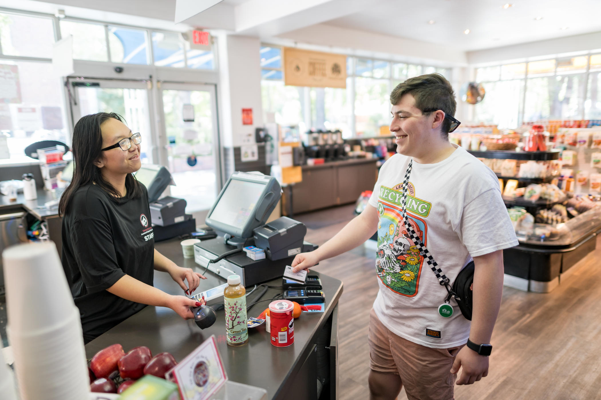 Jaylen Kruth (left) stands behind a counter as she accepts a purchase by Josh Rodriguez (right) who is using his EBT card to pay. There are a variety of snacks on the counter.