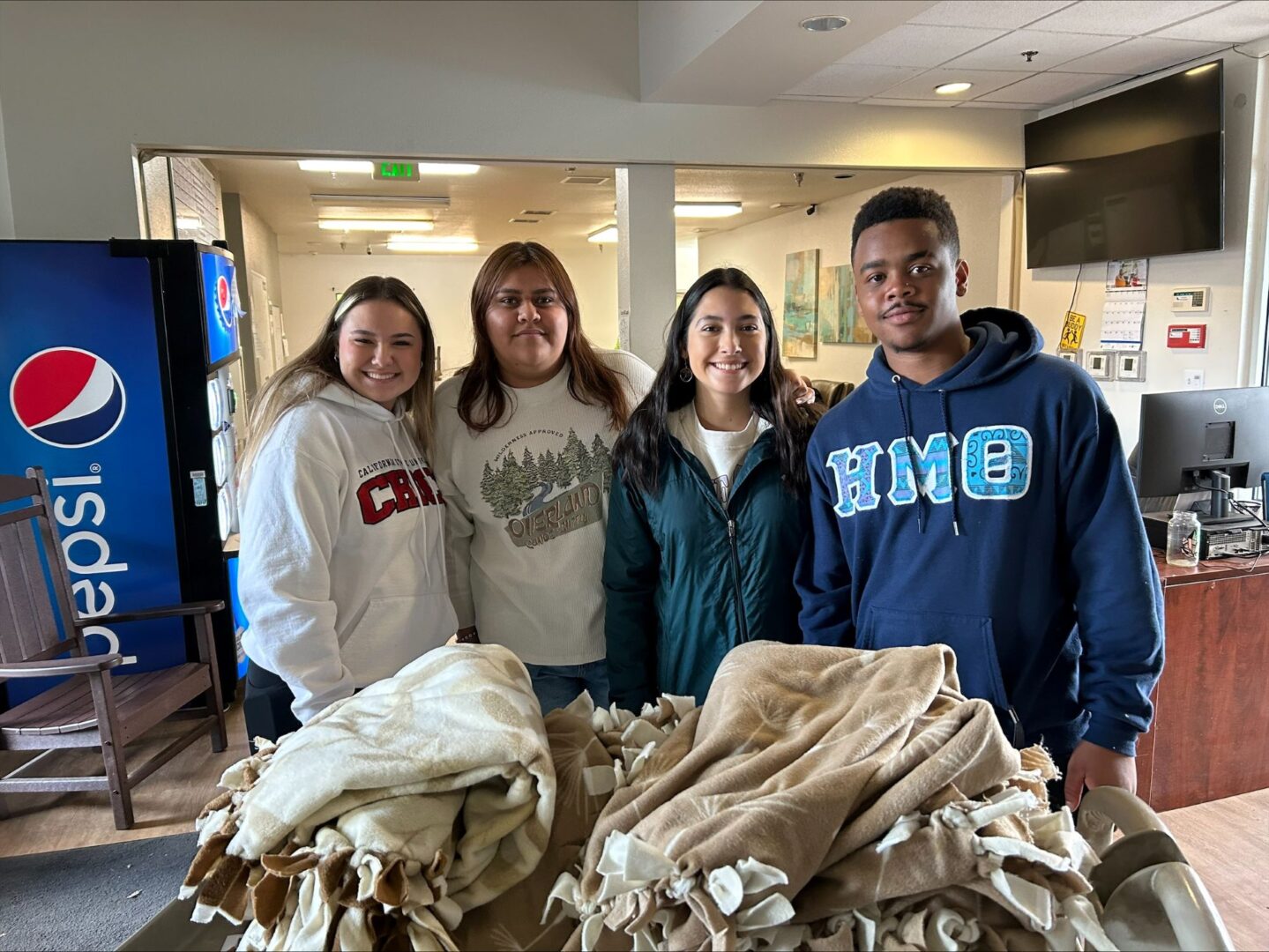 Four students stand shoulder to shoulder inside a room while a pile of blankets is on a table in front of them. 
