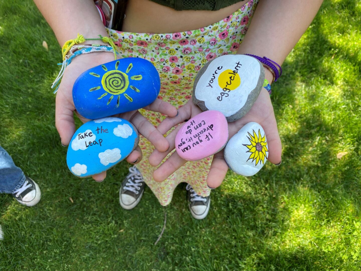 Two hands are holding five rocks that have been painted with positive messages, a sun, and one has a sunflower.