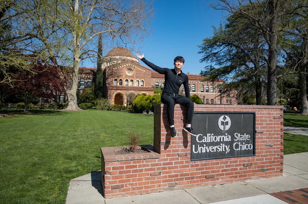 Haeryong Cheong sits atop the sign in front of Kendall Hall on campus, which says 'California State University, Chico'. He has his arm outstretched.