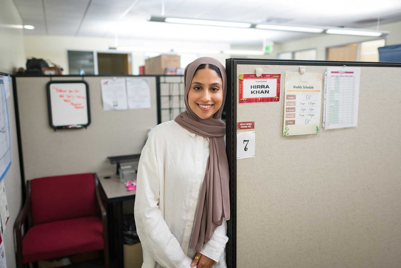Hirra Khan is photographed outside of her cubicle office at the Educational Opportunity Program. She wears a shite cotton shirt and a tan hijab. 