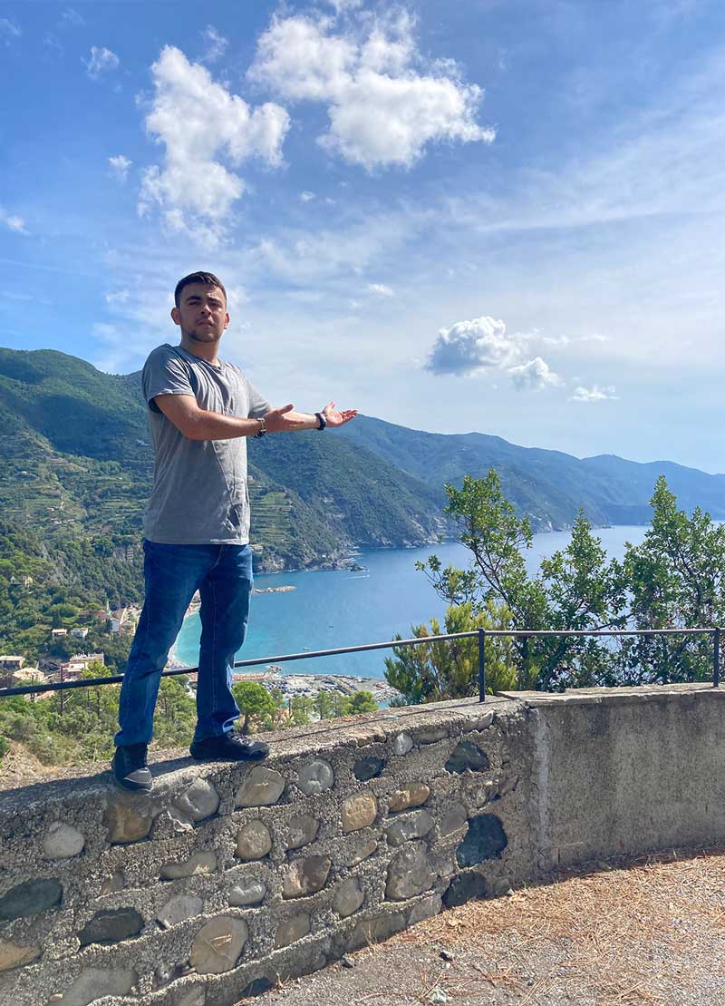 Humberto Partida stands on top of a small wall overlooking the powder-blue ocean in Italy. In the distance are green mountains are flanked by wispy clouds.